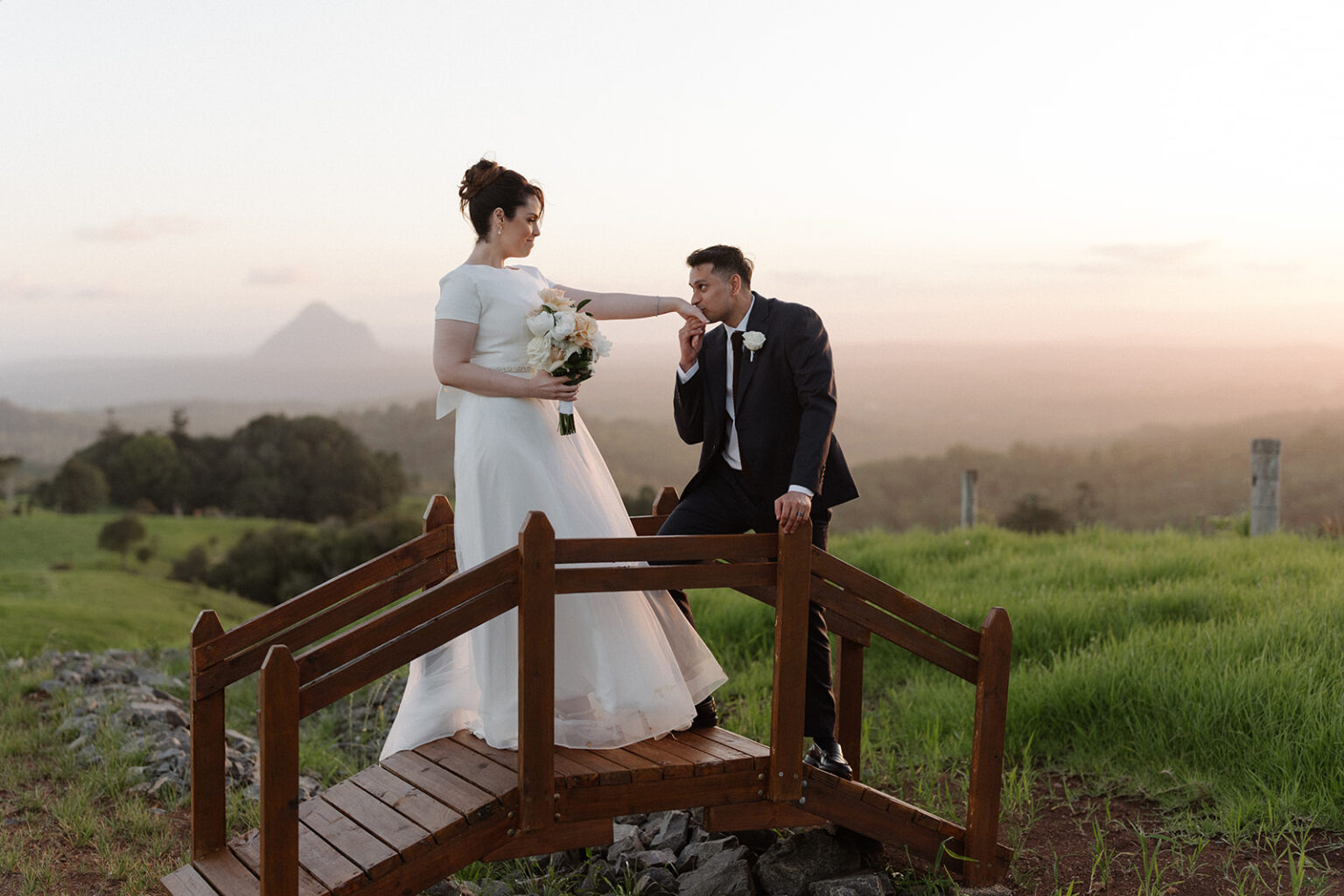 Groom kissing the bride's hand on a small timber bridge at sunset