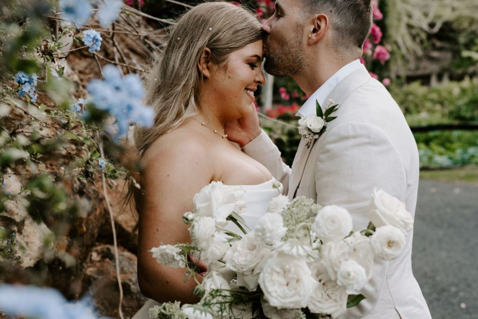 Groom kissing the bride's forehead beside a floral arrangement
